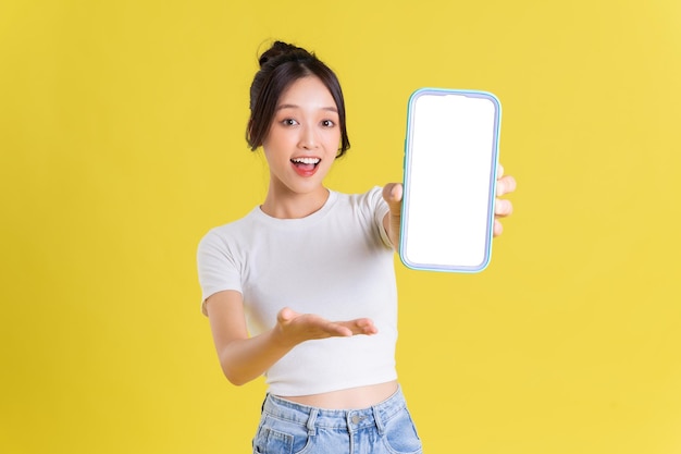 Young asian woman holding phone with cheerful face on yellow background