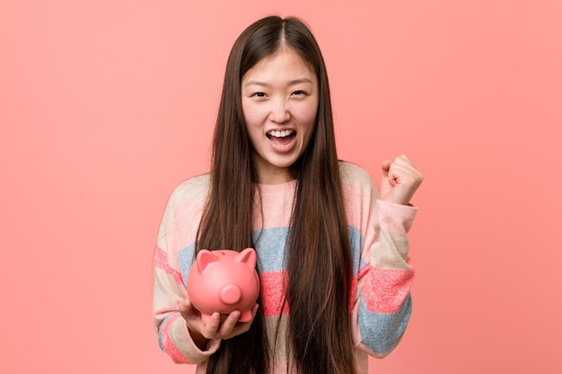 Young asian woman holding a piggy bank cheering carefree and excited. Victory concept.