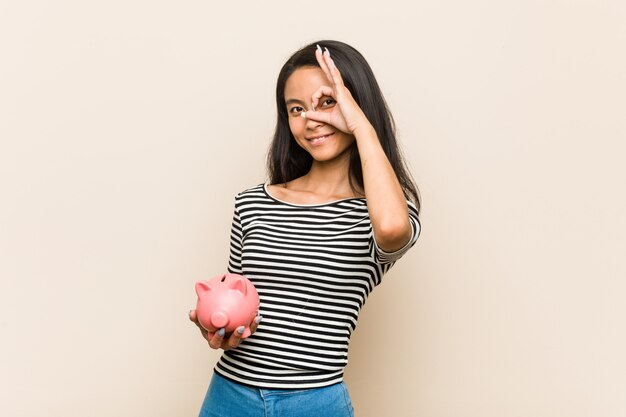 Young asian woman holding a piggy bank excited keeping ok gesture on eye.