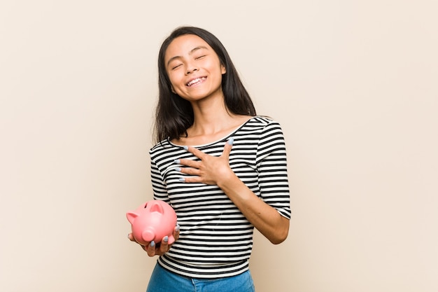 Young asian woman holding a piggy bank laughs out loudly keeping hand on chest.