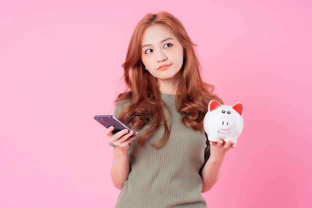 Young Asian woman holding piggy bank on pink background