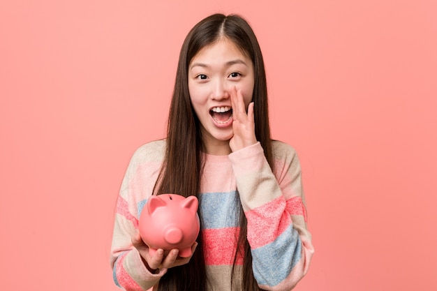 Young asian woman holding a piggy bank shouting excited to front.