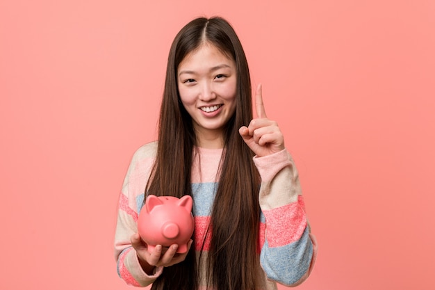 Young asian woman holding a piggy bank showing number one with finger.