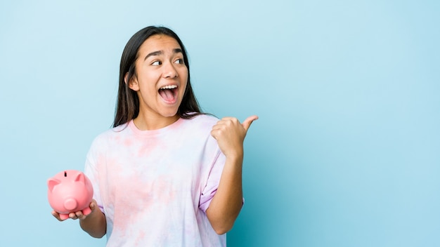 Young asian woman holding a pink bank over isolated background points with thumb finger away, laughing and carefree.