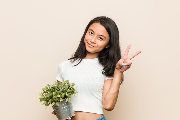 Young asian woman holding a plant joyful and carefree showing a peace symbol with fingers.