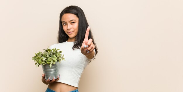 Young asian woman holding a plant showing number one with finger