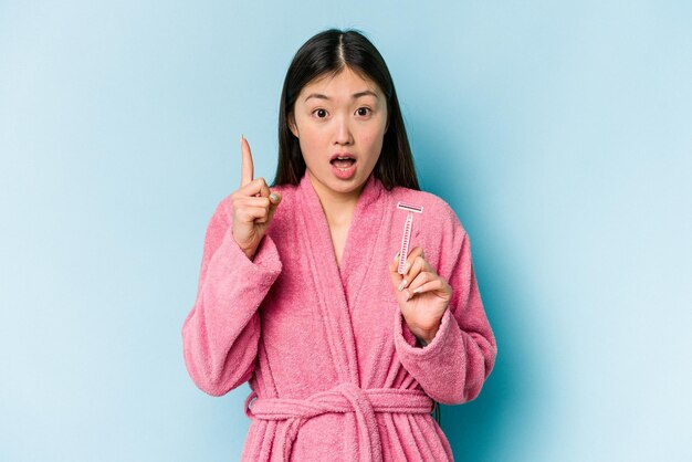 Young asian woman holding razor blade isolated on blue background having an idea inspiration concept