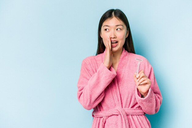 Young asian woman holding razor blade isolated on blue background is saying a secret hot braking news and looking aside