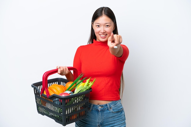 幸せな表情で正面を指している白い背景に分離された食品でいっぱいの買い物かごを保持している若いアジア女性