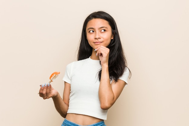 Young asian woman holding a sushi piece looking sideways with doubtful and skeptical expression.