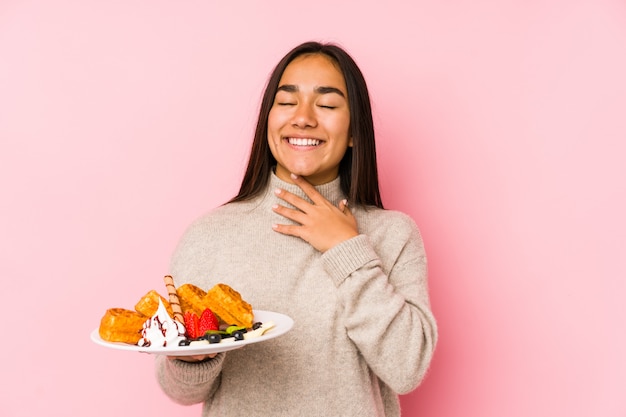 Young asian woman holding a waffle isolated laughs out loudly keeping hand on chest.