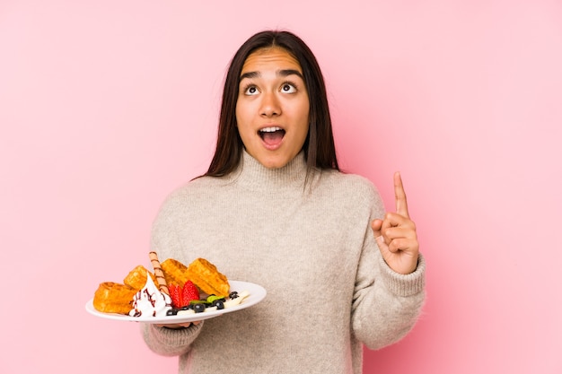 Young asian woman holding a waffle isolated pointing upside with opened mouth.