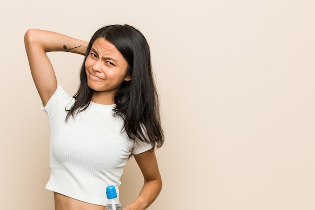 Young asian woman holding a water bottle touching back of head, thinking and making a choice.