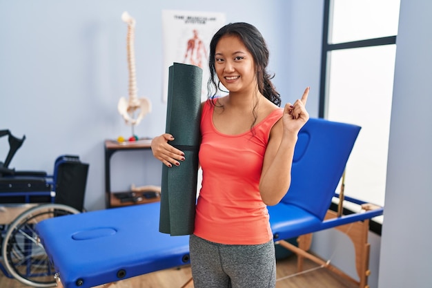 Young asian woman holding yoga mat at clinic smiling happy pointing with hand and finger to the side