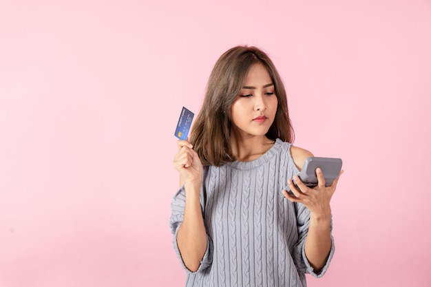 A young Asian woman holds a credit card and looks at the price of the product on the calculator. Isolated on pink background