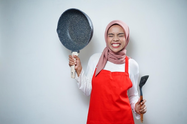 Молодая азиатская домохозяйка в кухонном фартуке готовит и держит сковороду и шпатель