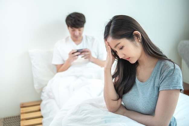 Young asian woman is upset that man uses phone all the time without paying attention to her. Man holds phone pay attention on smart mobile phone while sit on bed with ignoring on his girlfriend.