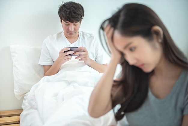 Young asian woman is upset that man uses phone all the time without paying attention to her. Man holds phone pay attention on smart mobile phone while sit on bed with ignoring on his girlfriend.