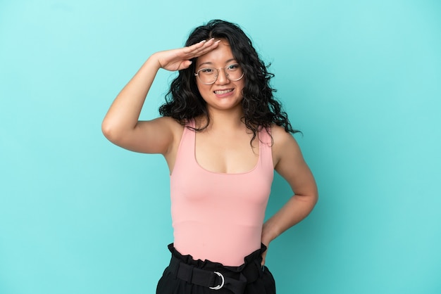 Young asian woman isolated on blue background saluting with hand with happy expression