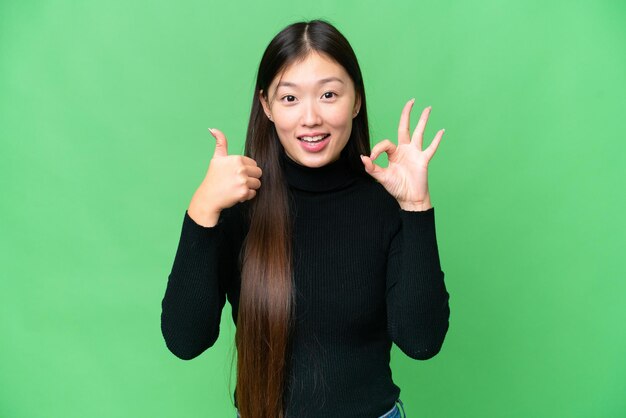 Молодая азиатка на изолированном фоне хроматического ключа показывает знак "ОК" и жест "большой палец вверх"
