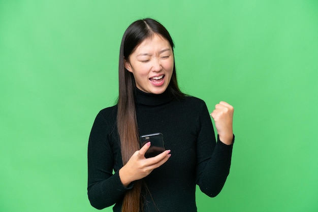 Giovane donna asiatica su sfondo chroma key isolato con il telefono in posizione di vittoria