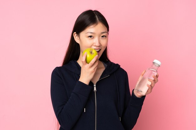 Young asian woman isolated on pink background with a bottle of water and eating an apple