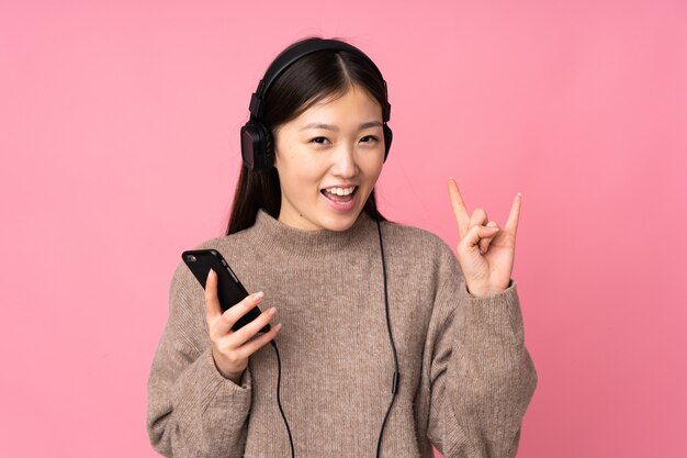 Young asian woman isolated on pink space listening music with a mobile making rock gesture