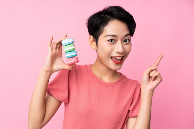 Young Asian woman over isolated pink wall holding colorful French macarons and pointing up a great idea