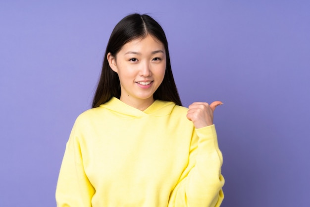 제품을 제시하기 위해 측면을 가리키는 보라색 배경에 고립 된 젊은 아시아 여자