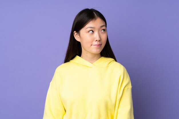 Young asian woman isolated on purple wall having doubts while looking up