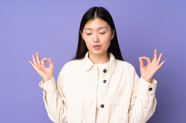 Young asian woman isolated on purple wall in zen pose