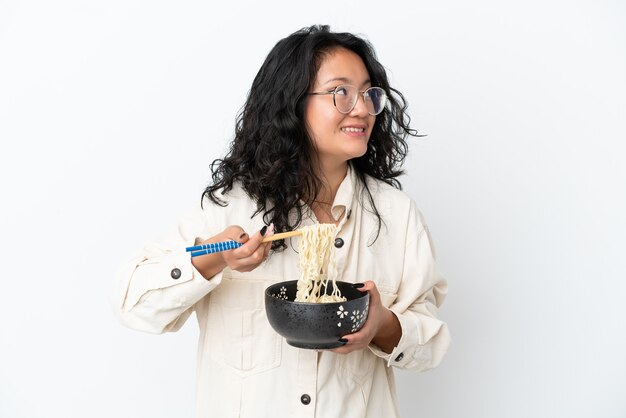 Молодая азиатская женщина, изолированная на белом фоне, держит миску лапши с палочками для еды и смотрит вверх