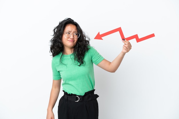 Young asian woman isolated on white background holding a downward arrow and with sad expression