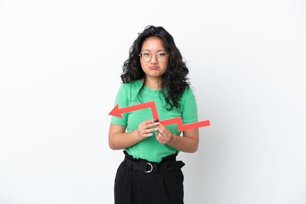 Young asian woman isolated on white background holding a downward arrow and with sad expression