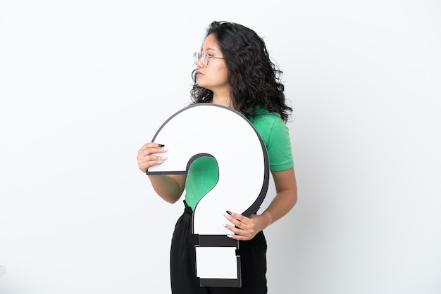 Young asian woman isolated on white background holding a question mark icon and looking side