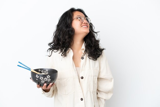 Young asian woman isolated on white background looking up while smiling while holding a bowl of noodles with chopsticks