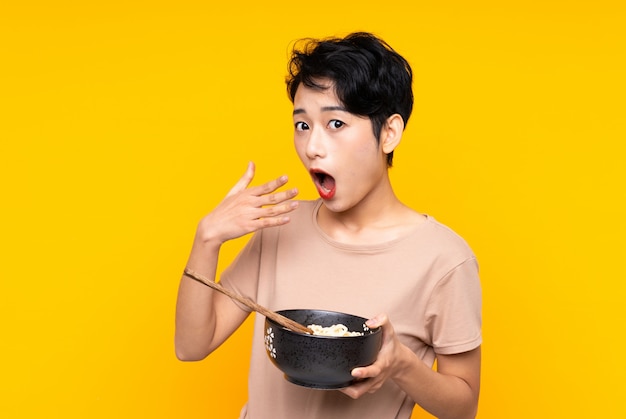 Young Asian woman over isolated yellow wall holding a bowl of noodles with chopsticks and surprised