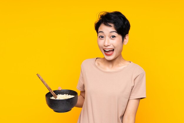 Young Asian woman over isolated yellow wall with surprise and shocked facial expression while holding a bowl of noodles with chopsticks