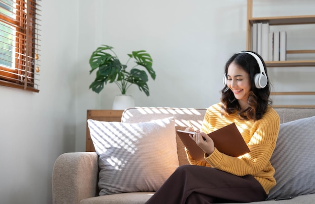 Молодая азиатская женщина слушает музыку в наушниках и пишет заметку о своей рабочей идее в дневнике. Она сидит на сером диване в гостиной