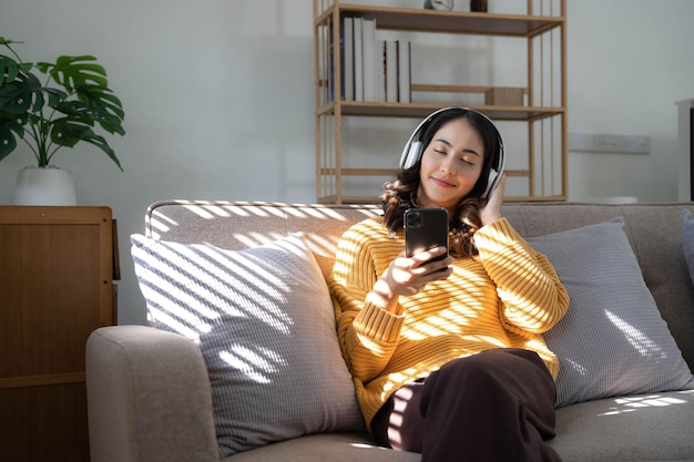 Young Asian woman listening music from headphones and writing note for her work idea She sitting on grey sofa in living room