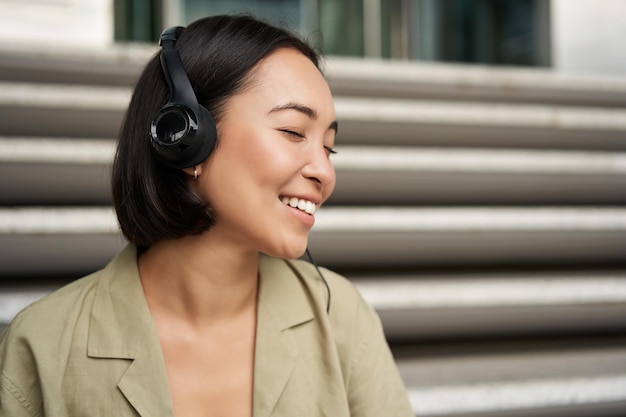 Young asian woman listening to music in headphones sitting outside on street smiling