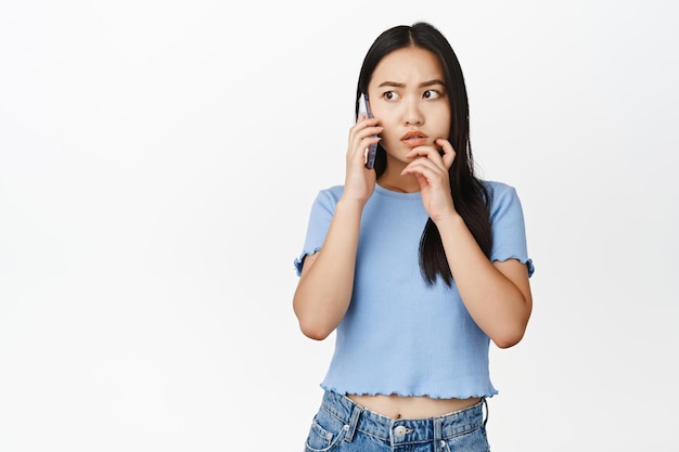 Young asian woman looking troubled during phone call anxious while talking on cellphone white background