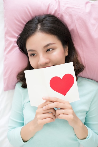 Young asian woman lying on bed, holding greeting card with red heart shape.