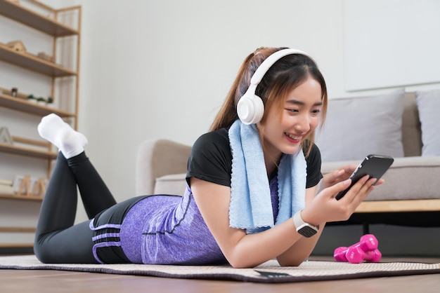 Young asian woman lying on floor to surfing internet and listens music on smartphone after exercise