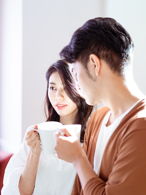 젊은 아시아 여성과 남성이 손에 커피 한 잔을 들고 집에서 함께 시간을 보내고 있습니다.