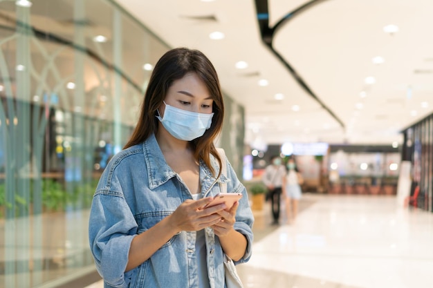 Young Asian woman in medical mask using mobile phone while walking at department store or public place New normal lifestyle health care concept