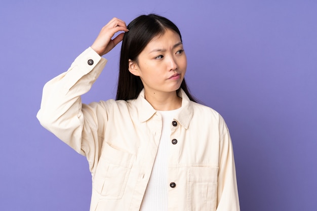 Young asian woman on purple wall having doubts while scratching head