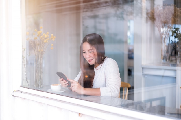 若いアジアの女性は、コーヒーショップのガラス窓から撮影した携帯電話を使用してリラックスします。