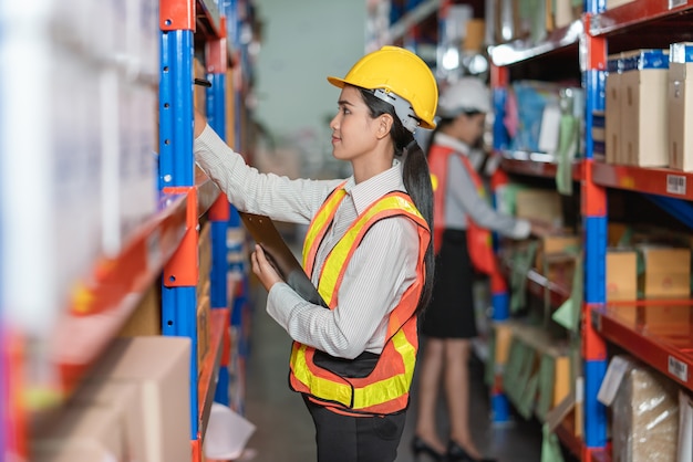 Young Asian woman in safety vest and helmet checking products in storage shelf