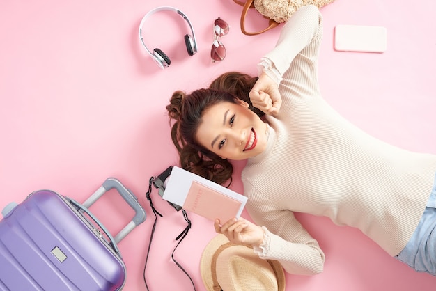 Young Asian woman showing her passport while lying on pink floor with travel luggage aurround